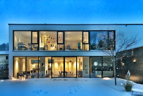 A modern house with large windows, snow in front, decorated with lights and visible interior rooms at dusk.